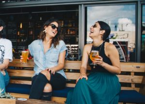 Two women sitting next to each other on a bench, holding glasses of beer and laughing.